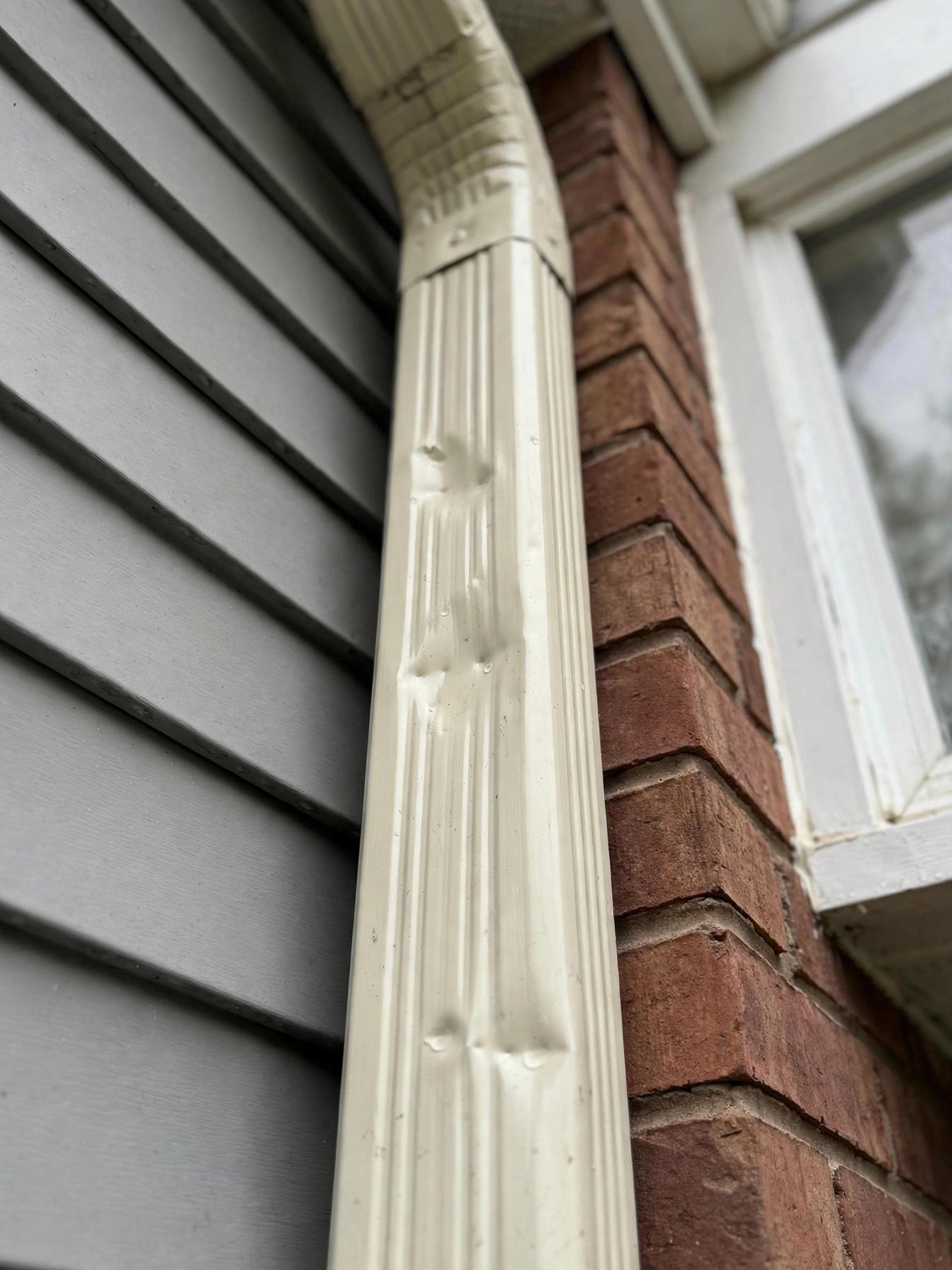Cream-colored downspout beside vinyl siding and brick with a few small hail dents on it